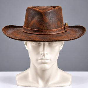 May include: A brown fedora hat with a distressed, weathered texture. The hat features a pinched crown, a wide brim, and a matching brown band around the base. A small, metallic brass-colored buckle is attached to the side of the hat band.