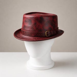 Può includere: Un cappello fedora con una finitura screziata rosso scuro e nera. Il cappello presenta una corona strutturata e una tesa, accentuata da una fascia abbinata rossa e nera con una piccola fibbia metallica color ottone sul lato esterno.