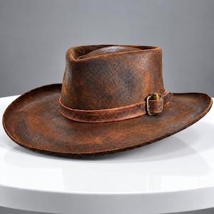 May include: A distressed brown hat with a wide brim and a high crown. The hat features a matching brown leather band wrapped around the base, secured by a metallic buckle with a small brass-coloured cylindrical accent attached to the side.