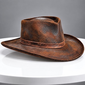 May include: A distressed brown fedora hat with a textured, weathered finish. The hat features a pinched crown and a wide, slightly curved brim, accented by a matching brown band encircling the base of the crown, creating a rugged appearance.