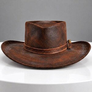 May include: A distressed brown cowboy hat with a weathered, textured finish. The hat features a wide, curved brim and a matching brown leather band around the crown, accented with a small, metallic, bullet-shaped decorative element on the side.