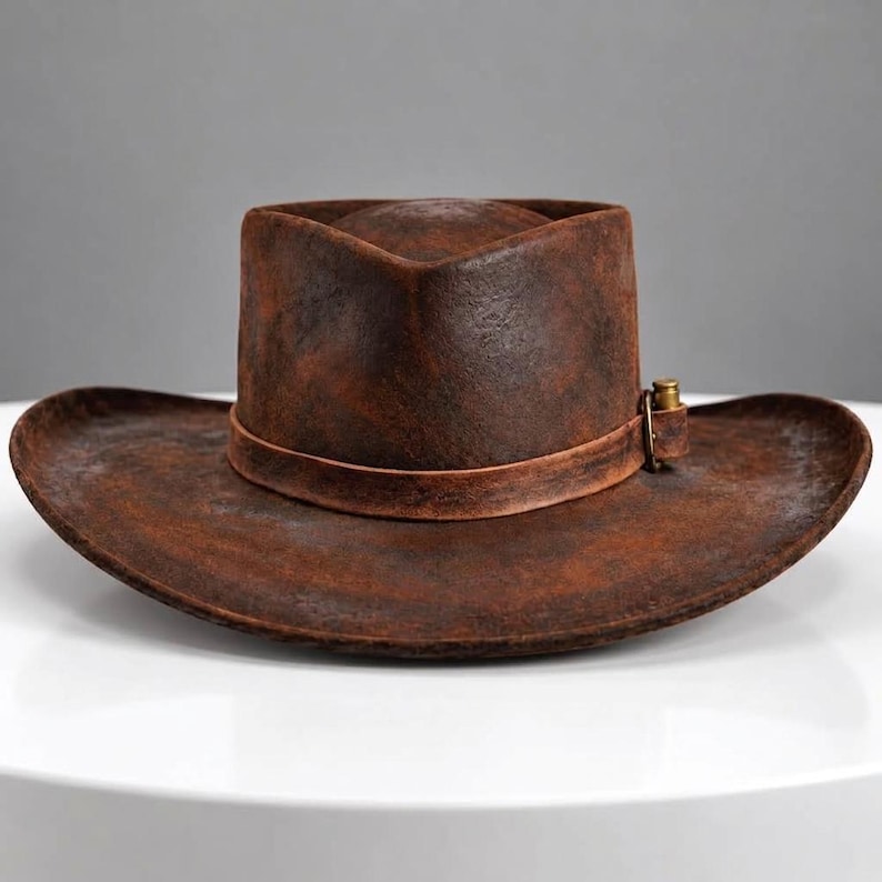 May include: A distressed brown cowboy hat with a weathered, textured finish. The hat features a wide, curved brim and a matching brown leather band around the crown, accented with a small, metallic, bullet-shaped decorative element on the side.