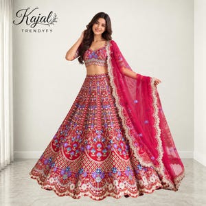 May include: A red lehenga choli set featuring intricate floral embroidery in blue, purple, and gold thread. The ensemble includes a cropped blouse, a voluminous floor-length skirt, and a matching sheer dupatta with scalloped edges. Text reads Kajal TRENDYFY.