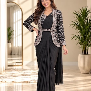 Puede incluir: Un sari negro combinado con una chaqueta de manga larga a juego con intrincados bordados florales en rosa y blanco. La chaqueta está ceñida a la cintura con un cinturón bordado a juego. El texto Kajal TRENDYFY es visible en la esquina superior.