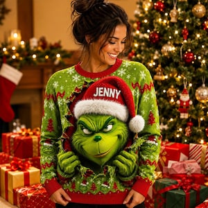 May include: A green long-sleeve jumper featuring a graphic of a green character wearing a red Santa hat with the name JENNY printed in white. The jumper has a pattern of white snowflakes and red Christmas trees with a ripped fabric effect.