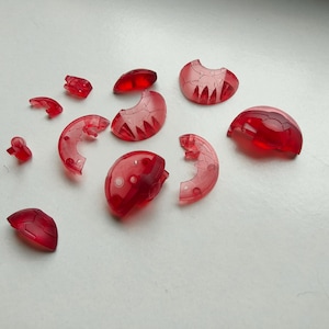 Peut inclure: Un ensemble de onze petits composants en plastique rouge translucide avec des lignes de panneaux géométriques moulées. Ces pièces individuelles varient en forme, incluant des formes courbes, semi-circulaires et angulaires.