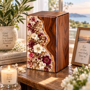 Pode incluir: Uma urna funerária retangular de madeira com um painel vertical de flores secas incrustadas em tons de vermelho escuro, rosa e branco. A madeira tem um veio castanho rico com uma curva natural que separa a secção floral da madeira maciça.