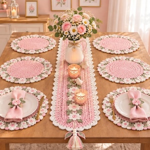 Op de afbeelding: Een tafelset bestaande uit een lange gehaakte tafelloper met roze en witte bloemmotieven, bijpassende ronde gehaakte placemats en roze stoffen servetten vastgezet met bloemvormige gehaakte servetringen voor een formele tafelsetting.