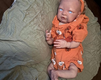 May include: A realistic baby doll with light skin and bright blue eyes, wearing an orange romper featuring a repeating black pumpkin print and a matching orange headband. The doll has light-coloured hair and is positioned on a soft olive green fabric.