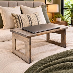 May include: A rectangular wooden bed tray with a dark brown, weathered wood grain finish and four sturdy legs. The tray features a flat, solid surface currently supporting a closed, thin, dark gray laptop computer centered on the top panel.