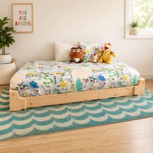 May include: A low-profile light wood bed frame with a white mattress cover featuring a colorful doodle pattern of musical notes, stars, and clouds. Three plush character toys sit on the bed, which is placed on a teal and white wave-patterned rug.