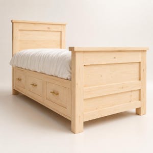 May include: A light-colored wooden bed frame featuring a tall headboard and a footboard with a horizontal plank design. The base includes three integrated storage drawers with small metallic handles, topped with a white quilted mattress cover.