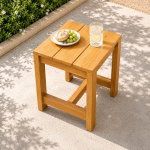 May include: A square wooden side table with a slatted top surface. On the table sits a white plate containing a round bread roll and a cluster of green grapes, alongside a clear glass filled with water and a slice of lemon.