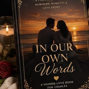 May include: A black hardback book titled In Our Own Words, A Guided Love Book For Couples. The cover features a sunset beach scene with a couple, gold decorative borders, and the text A Collection Of Our Memories, Moments & Love Story.