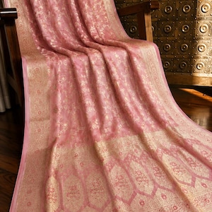 Puede incluir: Una tela drapeada de color rosa claro con un intrincado patrón floral metálico dorado en toda la superficie. La tela incluye un borde dorado ancho y ornamentado en la parte inferior con motivos de arco apuntado y bordados botánicos.