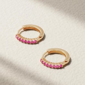 Puede incluir: Un par de pendientes de aro dorados con una hilera de pequeñas piedras circulares de color rosa vibrante engastadas a lo largo de la mitad frontal de cada aro. Los pendientes tienen un mecanismo de cierre de bisagra y un acabado metálico pulido.