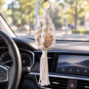 May include: A cream-colored macrame car air freshener holder featuring a knotted cord design, a circular wooden base, and a wooden sphere insert. The piece is suspended from a metal ring and finished with a long, fringed tassel at the bottom.