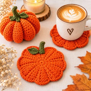 Pode incluir: Uma abóbora de crochê laranja com caule e folhas verdes, um porta-copos de abóbora de crochê plano e uma caneca de cerâmica com um ícone de coração contendo um latte com espuma. A caneca repousa sobre um segundo porta-copos de abóbora.