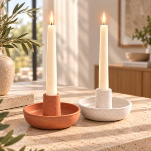 Puede incluir: Dos velas cónicas con llamas encendidas, cada una en un portavelas circular. Un soporte es de arcilla mate color terracota y el otro de piedra blanquecina moteada, ambos con un pilar central para sostener la vela.