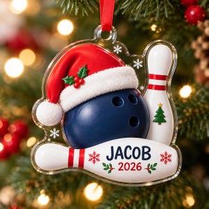 May include: A clear acrylic Christmas ornament featuring a dark blue bowling ball wearing a red and white Santa hat, next to a white bowling pin. The ornament includes the name JACOB and the year 2026 printed in blue and red with snowflake accents.