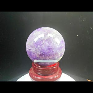 Puede incluir: Una esfera de cristal de amatista pulida con tonos púrpuras profundos y vetas blancas internas, que descansa sobre un soporte circular de madera de color marrón rojizo oscuro. La superficie de la piedra refleja la luz.