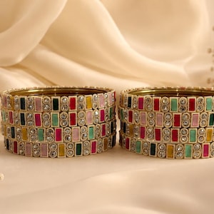 May include: Two stacks of circular bangles featuring alternating rectangular segments in red, pink, yellow, green, and light blue, interspersed with small clear circular stones set in a gold-toned metal frame with a smooth, polished finish.