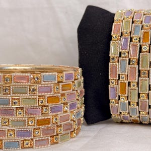 May include: A set of gold-toned bangles featuring rectangular enamel inlays in pastel shades of pink, blue, green, and orange. Each rectangular segment is bordered by small, clear, sparkling rhinestones set into the metallic frame.