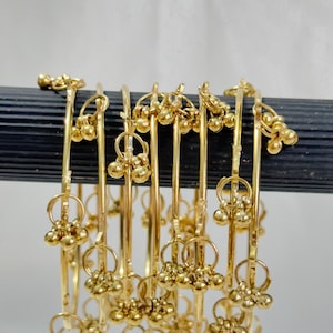 Könnte beinhalten: Ein Set aus goldfarbenen Metallarmreifen, die auf einer dunklen horizontalen Stange angeordnet sind. Jeder dünne, kreisförmige Armreif ist mit mehreren kleinen, kugelförmigen Anhängern versehen, die an winzigen Ringen befestigt sind.