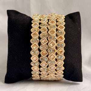 Puede incluir: Un conjunto de cuatro brazaletes dorados con un patrón repetitivo de elementos en forma de diamante y pequeños acentos circulares. Cada brazalete está construido con un acabado metálico texturizado e incrustado con piedras claras.