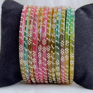 Könnte beinhalten: Ein Set aus durchscheinenden Armreifen in verschiedenen Farben wie Pink, Gelb, Grün und Blau. Jeder Armreif ist mit einem sich wiederholenden Muster aus kleinen, klaren, funkelnden Steinen in Rautenform auf der gesamten Oberfläche verziert.
