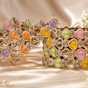Pode incluir: Um conjunto de pulseiras de metal em tom dourado com um padrão repetitivo de detalhes coloridos em forma de coração em tons de rosa, amarelo, laranja, verde e roxo, intercalados com cristais redondos transparentes e brilhantes.
