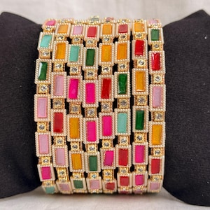 May include: A set of stacked bangles featuring rectangular, multi-colored enamel inlays in shades of pink, red, orange, green, and light blue. Each bangle is constructed with a gold-toned metal frame accented by small, clear, round rhinestones.