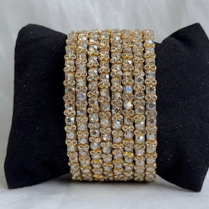 May include: A set of eight gold-toned bangles arranged in a stack. Each bangle features a repeating pattern of small, clear, faceted circular stones set in gold-coloured metal, creating a textured, reflective surface across the entire collection.