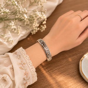 Könnte beinhalten: Ein silberfarbenes Armband mit einem komplizierten, durchbrochenen Filigranmuster aus geschwungenen Metallverzierungen. Das Armband wird am Handgelenk getragen und durch einen dünnen, zierlichen Ring mit kleinen Steinen am Finger ergänzt.