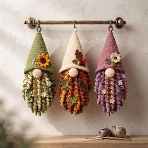 Peut inclure: Trois ornements de gnomes au crochet suspendus à une tringle en métal. Le gnome de gauche est vert avec un tournesol, celui du centre est crème avec des feuilles d'automne et des baies, et celui de droite est mauve avec des petites fleurs.
