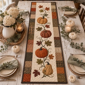 May include: A rectangular quilted table runner featuring a central cream panel with an embroidered vine of four pumpkins and autumn leaves, bordered by a patchwork of brown, orange, and green fabric squares, displayed on a rustic wooden table.