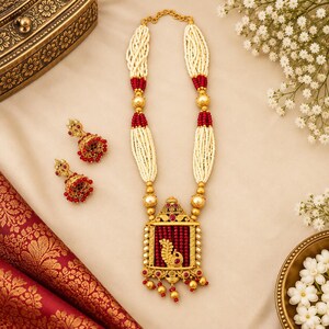 May include: A gold-toned necklace and matching pair of earrings featuring white seed bead strands, deep red beads, and a rectangular pendant with a central peacock motif. The necklace includes gold-toned spacers and small red bead accents throughout.