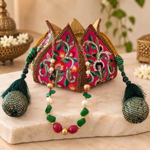 May include: A fuchsia drawstring pouch featuring intricate floral embroidery, sequins, and gold beadwork, finished with two large dark green tassels. A beaded necklace with green, red, and white beads rests in front of the pouch.