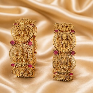 May include: Two gold-toned bangles featuring intricate embossed figures of deities within circular filigree frames. The bangles are accented with small, round, vibrant pink stones set between the circular motifs, creating a detailed metallic design.