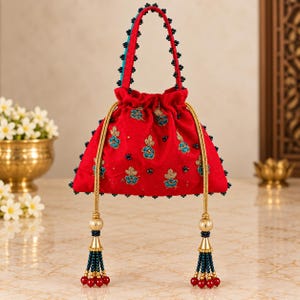 May include: A red drawstring handbag featuring embroidered blue and gold floral motifs, accented with dark blue bead trim along the edges and handle. The bag includes two long gold braided cords ending in decorative tassels with beads.