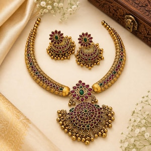 May include: A gold-toned necklace and matching jhumka earring set featuring intricate metalwork, embedded with deep red and green stones. The necklace has a central floral pendant with dangling gold beads, while the earrings mirror the design.