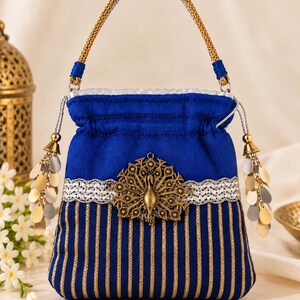 May include: A royal blue drawstring handbag featuring a gold metallic peacock emblem centered on a silver woven band. The lower section has vertical gold stripes, and the bag includes a braided gold handle with dangling metallic leaf-shaped charms.
