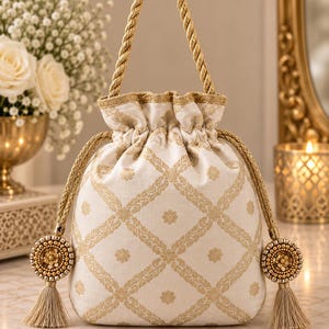 May include: A cream-colored drawstring pouch bag featuring a gold-toned lattice pattern with floral motifs. The bag includes a braided gold rope handle, gold drawstring cords, and two circular decorative tassels adorned with gold beads and sequins.