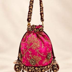 May include: A vibrant fuchsia drawstring pouch bag featuring intricate gold thread embroidery in floral patterns. The bag includes a dark maroon velvet handle and base trim, accented with decorative beaded tassels and hanging dark crystal beads.