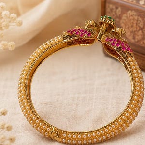May include: A gold-toned hinged bangle bracelet featuring a circular band encrusted with small white pearls. The clasp area is adorned with two stylized bird motifs embellished with vibrant pink and green stones, creating a detailed focal point.