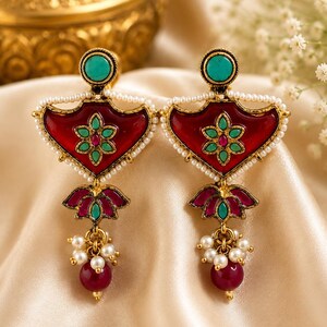 May include: A pair of gold-tone drop earrings featuring a shield-shaped red enamel center with a central green and pink floral motif. The edges are lined with small white beads, and each earring includes a dangling lotus-shaped charm with pearls.
