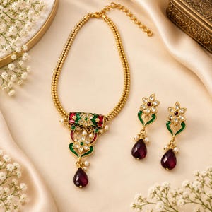 May include: A gold-toned necklace and matching drop earring set featuring green and red enamel detailing, clear stones, and small white pearls. The necklace has a double-strand chain and a central pendant with a deep red teardrop-shaped stone.