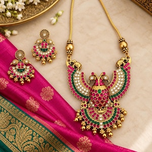 May include: A gold-toned necklace and matching earrings set featuring a central peacock motif. The jewelry is adorned with inlaid pink and green stones, small white crystals, and dangling gold beads. The necklace includes two small elephant figures.