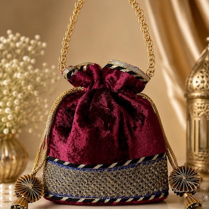 May include: A deep burgundy velvet drawstring handbag featuring a gold chain handle and a wide base band decorated with intricate gold embroidery, sequins, and blue trim. The bag includes two gold cord drawstrings finished with pleated gold tassels.