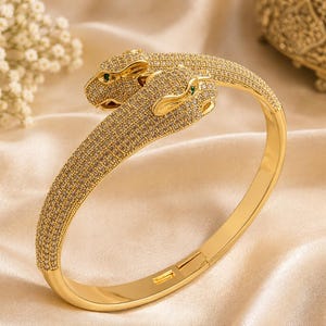May include: A gold-toned hinged bangle bracelet featuring two panther heads facing each other. The entire surface of the panther heads and the band is encrusted with small clear pavé stones, with two small green stones serving as the panther eyes.